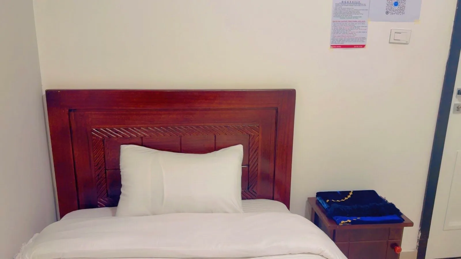 Single room with a carved mahogany headboard, one pillow on a white bed and a wooden bedside table